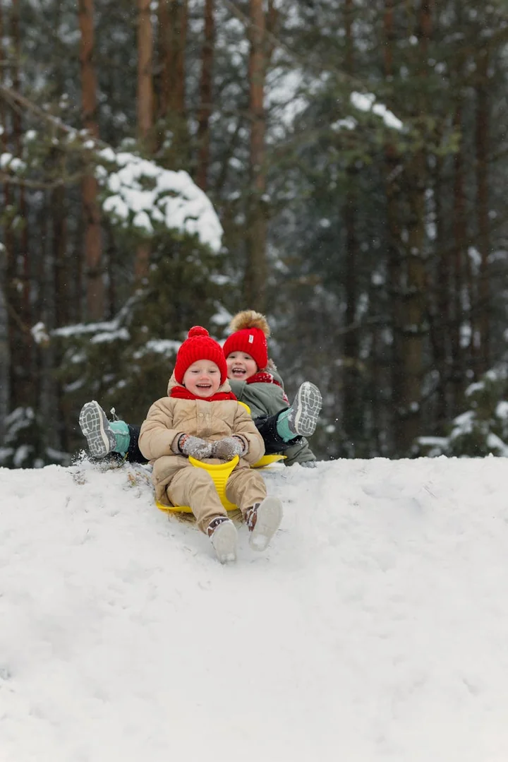 Dwoje uśmiechniętych dzieci w czerwonych czapkach zjeżdża na sankach po zaśnieżonym stoku.