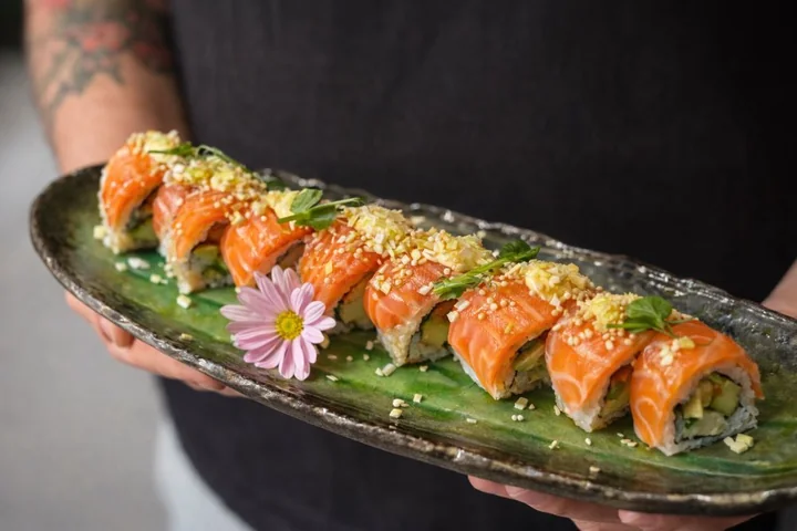 Talerz z ośmioma rolkami sushi z łososiem, awokado, posypką, mikroliśćmi i różowym kwiatkiem, trzymany w dłoniach