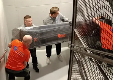 Czterech mężczyzn wnosi duży, owinięty folią przedmiot po schodach.