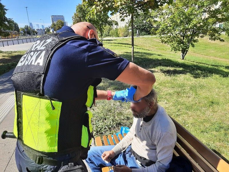 Strażnik miejski zakładający opatrunek na głowę bezdomnemu siedzącemu na ławce.