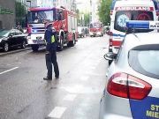 Zdjęcie ilustracyjne: strażnik miejski zabezpieczający ulicę. Na pierwszym planie fragment radiowozu straży miejskiej, w tle karetka, a w lewym górnym rogu- wóz strażacki.