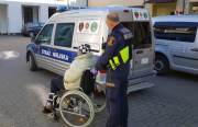 Zdjęcie z interwencji: strażnik miejski pchający wózek inwalidzki z siedzącym na nim bezdomnym. W tle stojący radiowóz straży miejskiej oraz pojazd Caritasu.