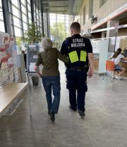 Strażnik miejski prowadzący pod ramię starszą kobietę na korytarzu urzędu dzielnicy w Wilanowie.