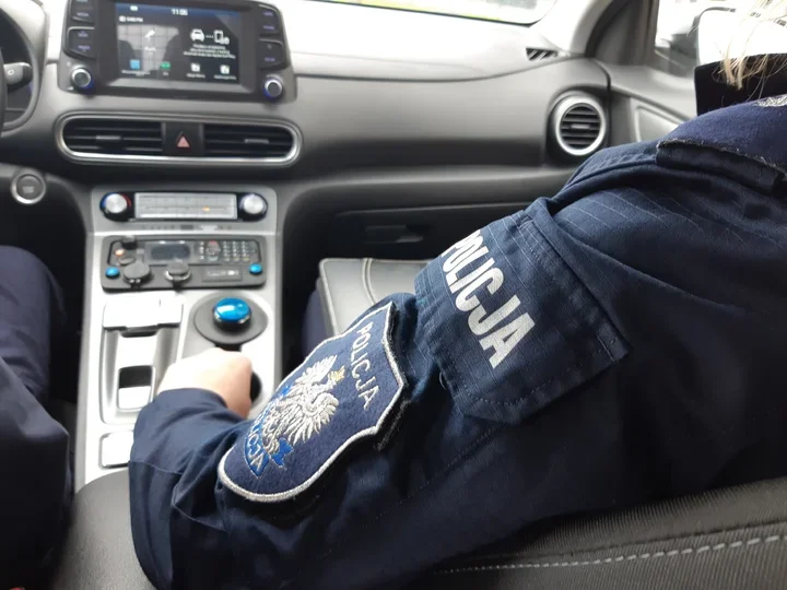 Funkcjonariusz policji w mundurze z napisem „POLICJA” siedzi na przednim siedzeniu radiowozu, trzymając rękę na konsoli środkowej z radiotelefonem.
