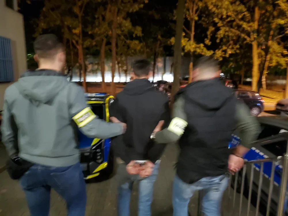 Funkcjonariusze w opaskach na rękawach prowadzą zatrzymanego w kajdankach mężczyznę do radiowozu nocą na ulicy.