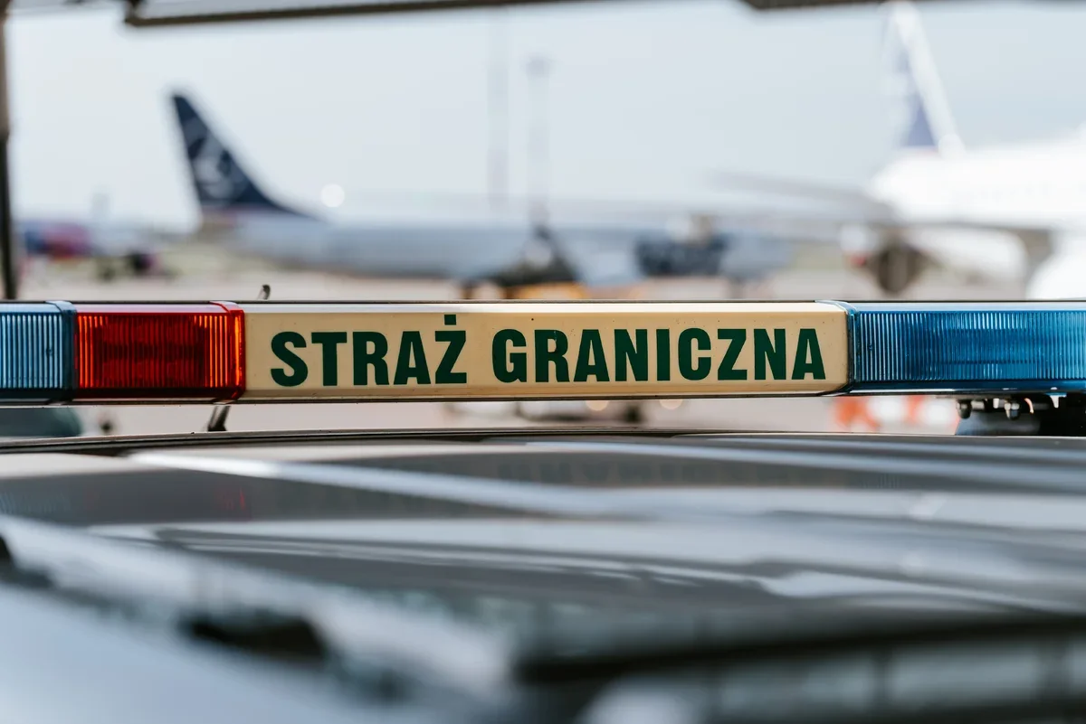 Lampa sygnalizacyjna radiowozu Straży Granicznej na lotnisku, w tle rozmyte samoloty na płycie postojowej.