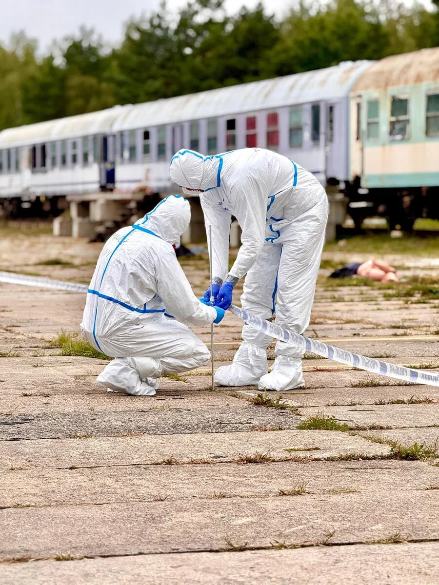Dwie osoby w kombinezonach ochronnych zabezpieczają teren taśmą policyjną na opuszczonej stacji kolejowej.
