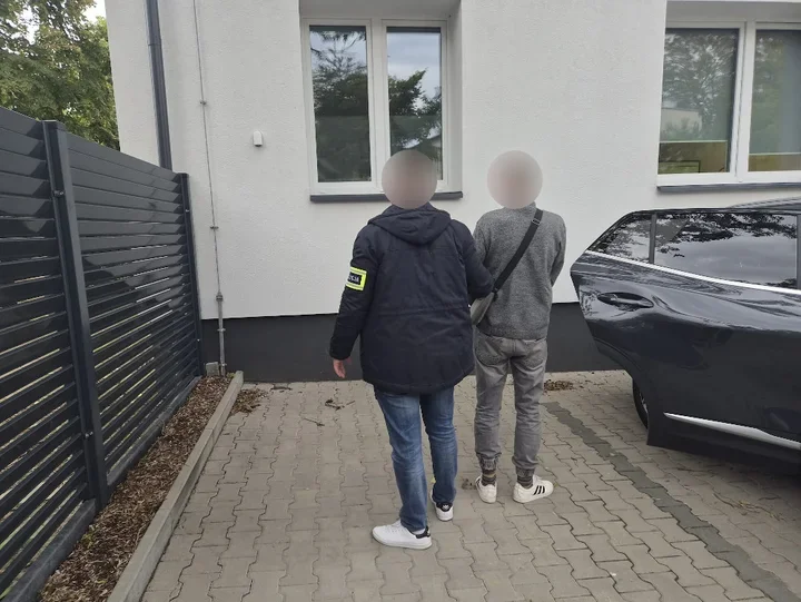 Funkcjonariusz w kurtce z żółtą opaską na rękawie prowadzi mężczyznę w szarej bluzie w stronę budynku przy zaparkowanym czarnym samochodzie.