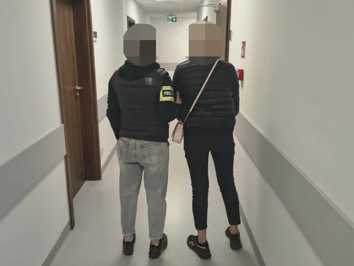 Funkcjonariusz w kamizelce z napisem POLICJA prowadzi kobietę korytarzem budynku użyteczności publicznej.