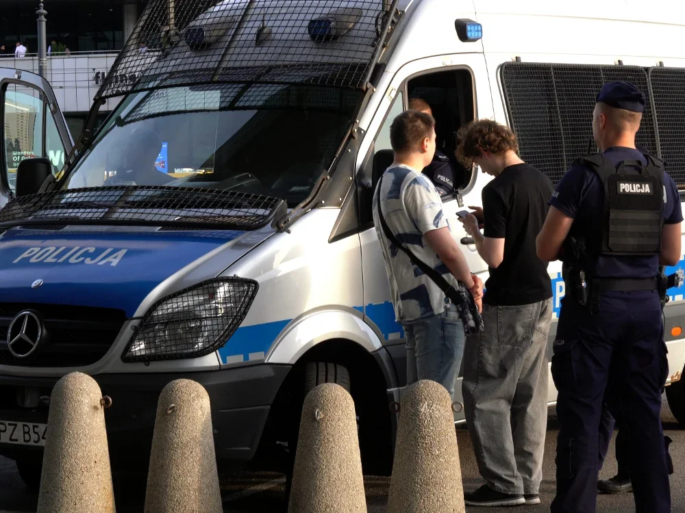 Policjanci przy policyjnym busie zabezpieczonym kratami prowadzą czynności z dwoma młodymi mężczyznami na ulicy.