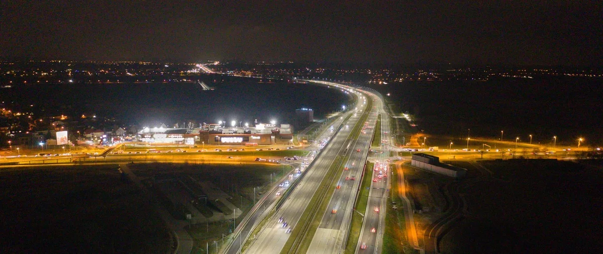 Nocny widok z lotu ptaka na oświetloną wielopasmową autostradę z ruchem samochodowym, węzeł drogowy oraz zabudowania na przedmieściach dużego miasta.