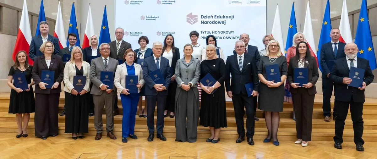 Grupa osób z dyplomami pozuje do zdjęcia podczas uroczystości Dnia Edukacji Narodowej z napisem „Tytuł Honorowy Przodownika Oświaty” na tle.