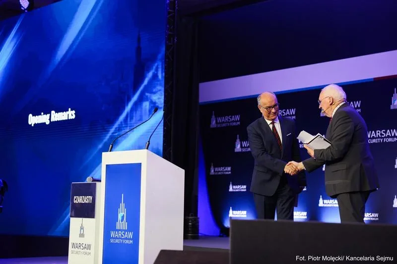 Dwaj mężczyźni w garniturach witają się uściskiem dłoni na scenie podczas Warsaw Security Forum, obok pulpit z napisem „Czarzasty”.