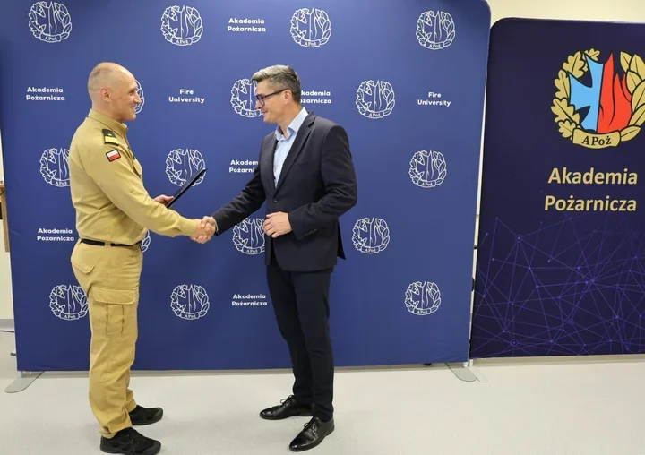 W pomieszczeniu przed ścianką Akademii Pożarniczej funkcjonariusz PSP w mundurze uroczyście wręcza dokument mężczyźnie w garniturze, który przyjmuje go uściskie