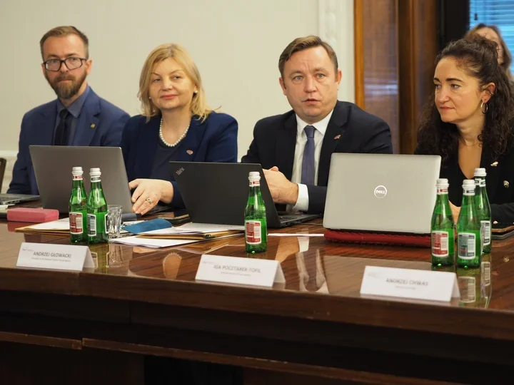 Cztery osoby siedzą przy drewnianym stole konferencyjnym z laptopami i butelkami wody, uczestnicząc w spotkaniu formalnym.