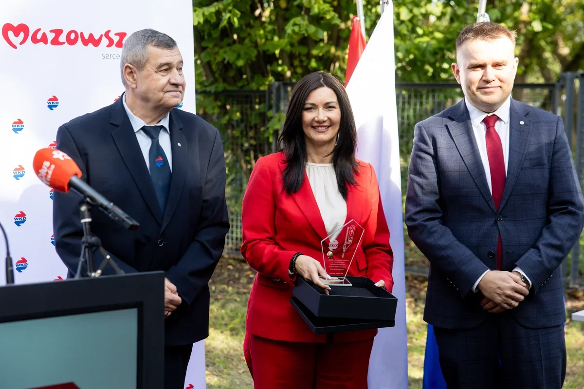 Trzy osoby w strojach biznesowych stoją na zewnątrz podczas uroczystości; kobieta w czerwonym garniturze trzyma szklaną nagrodę w czarnym pudełku, obok mównicy