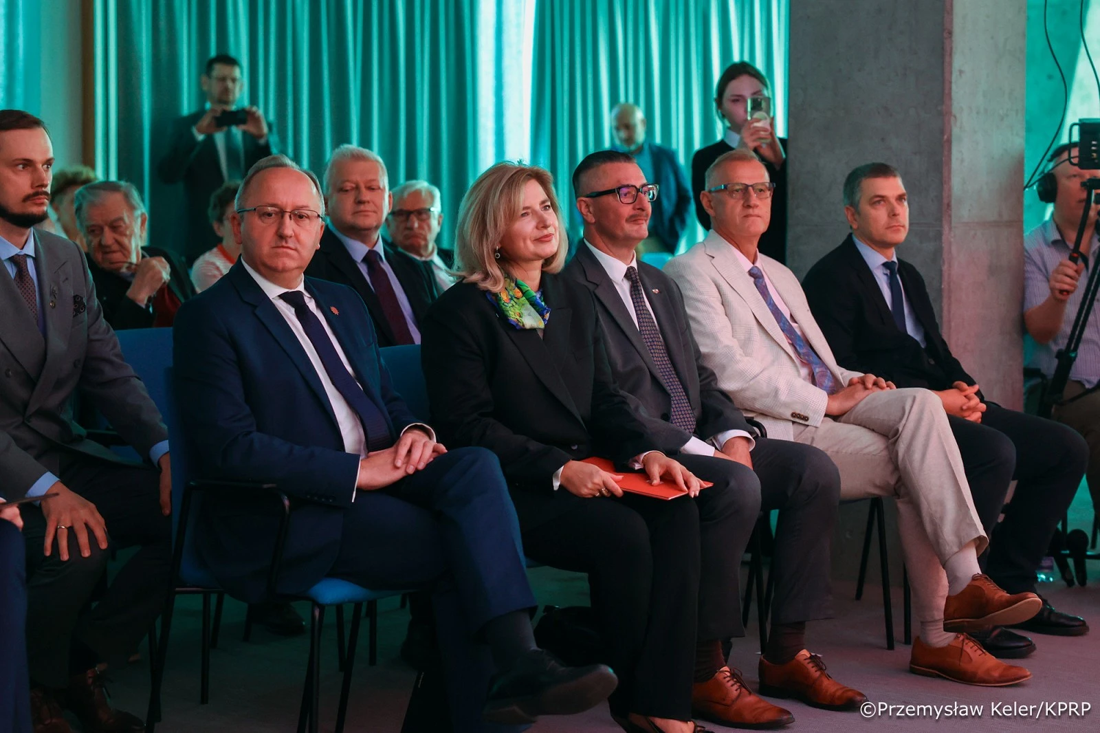 Uczestnicy oficjalnej uroczystości siedzą w pierwszym rzędzie sali oświetlonej zielonym światłem, w tle fotograf i kamerzysta.