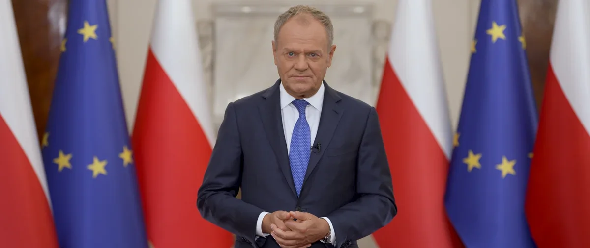 Mężczyzna w garniturze stoi przed flagami Polski i Unii Europejskiej w reprezentacyjnym wnętrzu.