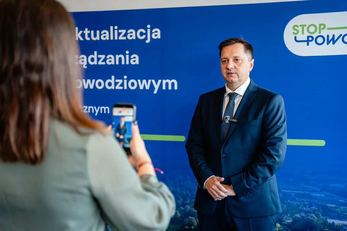 Mężczyzna w garniturze stoi przed niebieską ścianką z napisami dotyczącymi powodzi, a kobieta nagrywa go smartfonem.