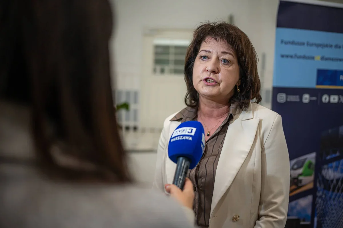 Kobieta w beżowej marynarce udziela wywiadu dziennikarce z mikrofonem TVP3 Warszawa podczas wydarzenia związanego z Funduszami Europejskimi.