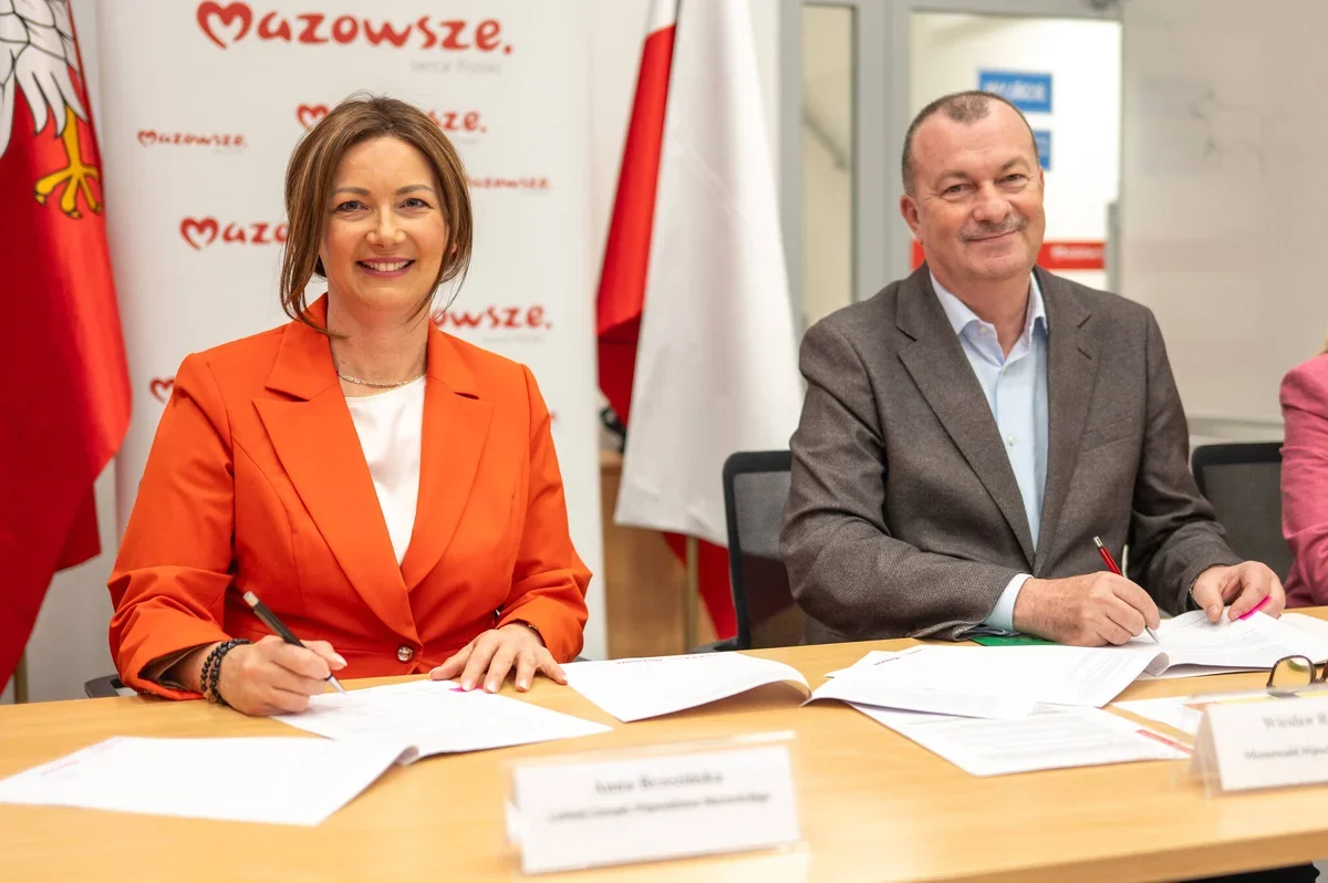 Dwie osoby siedzą przy stole i podpisują dokumenty podczas uroczystości w siedzibie samorządu województwa mazowieckiego.