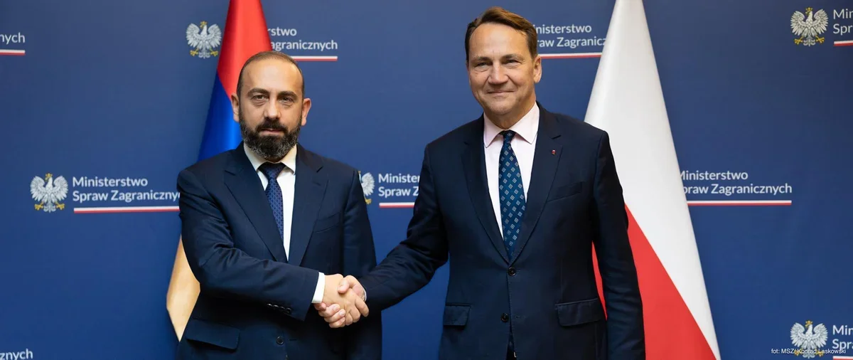 Dwaj mężczyźni w garniturach witają się uściskiem dłoni podczas spotkania w Ministerstwie Spraw Zagranicznych, w tle flagi Armenii i Polski.