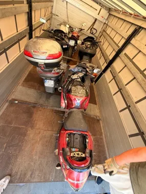Cztery motocykle stoją wewnątrz naczepy ciężarówki, jeden z nich ma otwarte siedzenie i jest przytrzymywany przez osobę w rękawicach.