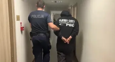Funkcjonariusz policji prowadzi zakuwanego w kajdankach mężczyznę korytarzem budynku.
