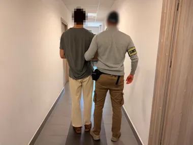 Funkcjonariusz policji prowadzi zatrzymanego mężczyznę po korytarzu budynku.