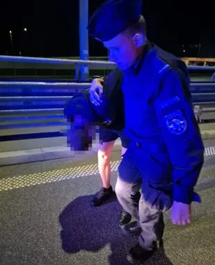 Funkcjonariusz służb mundurowych wspiera mężczyznę pochylonego przy barierze na moście nocą.