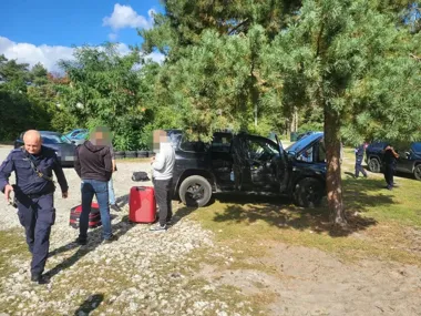 Funkcjonariusz policji i kilka osób stoją przy czarnym SUV-ie z otwartymi drzwiami na zalesionym parkingu, obok leżą walizki.