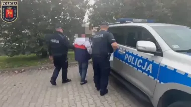 Dwóch policjantów prowadzi zatrzymaną osobę do policyjnego radiowozu zaparkowanego na brukowanej nawierzchni w otoczeniu zieleni.