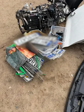 Na ziemi leżą stosy tablic rejestracyjnych, części samochodowe i plastikowe pojemniki.