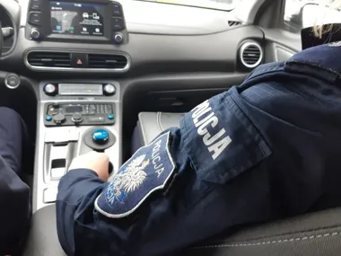 Funkcjonariusz policji w mundurze z napisem „POLICJA” siedzi na przednim siedzeniu radiowozu, trzymając rękę na konsoli środkowej z radiotelefonem.