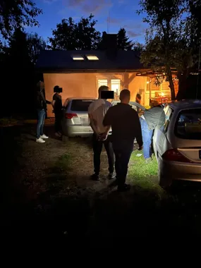 W nocy przed podświetlonym domem stoją i poruszają się kilka osób, w tym mężczyzna prowadzony z rękami złożonymi za plecami, przy zaparkowanych samochodach.