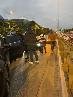 Policjanci przy czarnym samochodzie na moście o zmierzchu, ruch uliczny w tle.