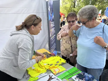 Kobieta rozdaje materiały promocyjne i odblaskowe dwóm starszym kobietom przy stoisku podczas plenerowej imprezy.