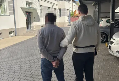 Funkcjonariusz prowadzi zatrzymanego mężczyznę z rękoma skutymi kajdankami na brukowanej ulicy między budynkami.