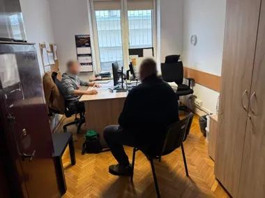 W biurowym pomieszczeniu z oknami dwie osoby siedzą przy biurku z komputerami, jedna z nich pracuje przy klawiaturze, druga siedzi naprzeciwko na krześle.