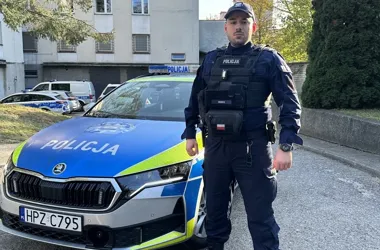 Policjant w mundurze stoi przed policyjnym radiowozem marki Škoda na ulicy przed budynkiem.