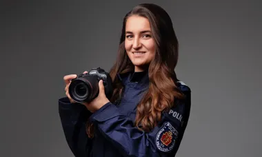 Kobieta w granatowej bluzie policyjnej trzyma w dłoniach aparat fotograficzny Canon EOS R5 na szarym tle.