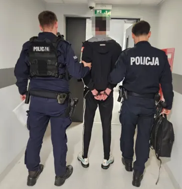 Dwóch policjantów w mundurach prowadzi zatrzymanego mężczyznę w kajdankach korytarzem budynku.