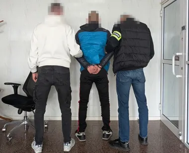 Policjant prowadzi zatrzymanego mężczyznę w kajdankach w pomieszczeniu biurowym.