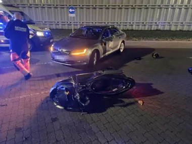 Nocna scena wypadku drogowego z udziałem samochodu osobowego i uszkodzonego skutera na brukowanej jezdni, przy obecności ratownika medycznego i karetki.