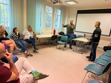 Dwóch mężczyzn prowadzi dyskusję lub prezentację przed grupą osób siedzących w sali konferencyjnej z ekranem projekcyjnym.