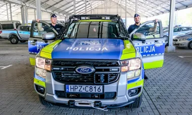 Dwaj policjanci stoją przy policyjnym pojeździe Ford Ranger oznaczonym „POLICJA” na parkingu pod wiatą.