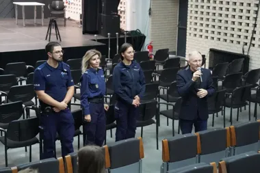 Trójka policjantów w mundurach i mężczyzna w garniturze z mikrofonem stoją w sali audytoryjnej przed rzędami krzeseł.