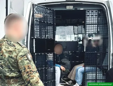 Funkcjonariusz Straży Granicznej stoi przy otwartym transporcie więziennym, w którym za kratami siedzą dwie osoby.