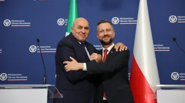 Dwaj mężczyźni w garniturach stoją na tle logotypu Ministerstwa Obrony Narodowej, uściskają sobie dłonie i obejmują się ramionami między flagami Włoch i Polski.