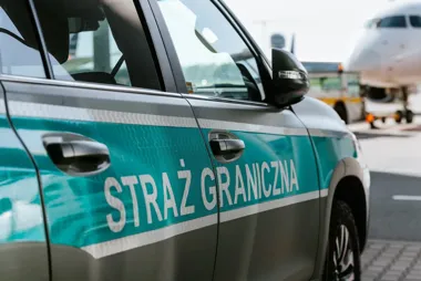 Samochód Straży Granicznej stoi na płycie lotniska, w tle widać samolot i pojazd obsługi naziemnej.